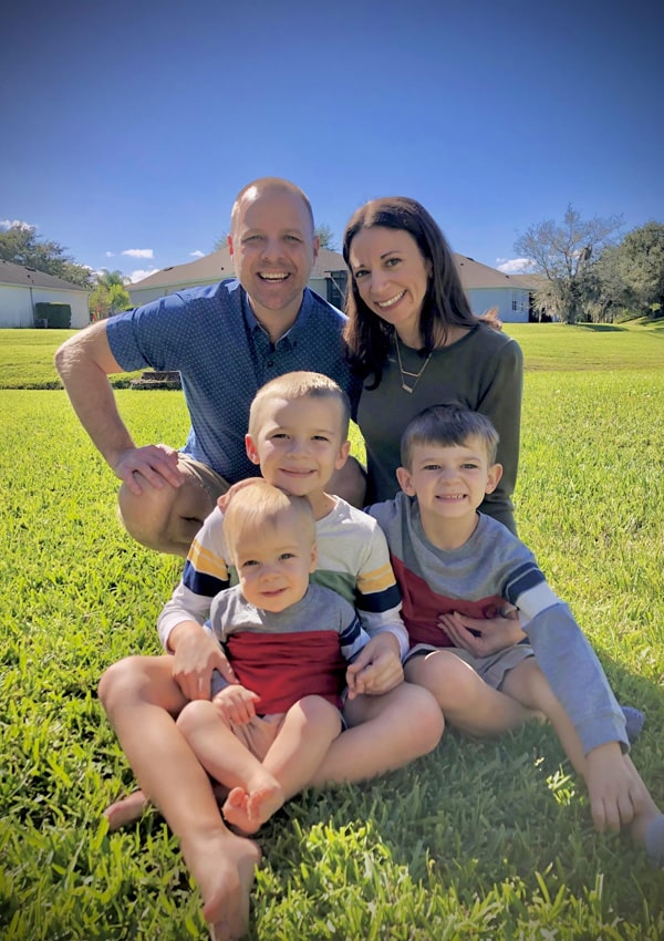 A smiling family of five, including a local vet, poses on green grass under a bright blue sky. Two adults kneel behind three young boys in casual clothes, with houses and trees visible in the background.