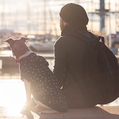 A veterinarian wearing a backpack and a beanie sits beside a dog in a patterned shirt, both facing a marina with boats at sunset.