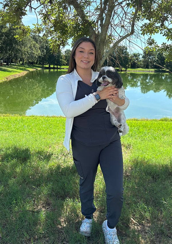 A woman, likely a veterinarian, stands on grass near a lake, smiling and holding a small black and white dog. She wears dark scrubs and a white jacket. Trees and the lake are in the background on a sunny day.