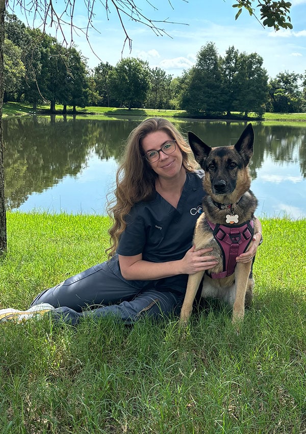 A woman in dark scrubs, likely a vet, sits on grass by a pond, smiling and holding a German Shepherd in a pink harness. Trees and blue sky form the background.