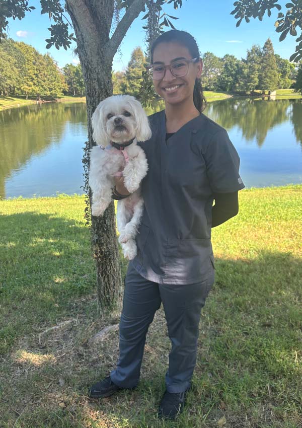 A smiling veterinarian in gray scrubs stands on grass by a lake, holding a small white dog. Trees and a blue sky brighten the background.